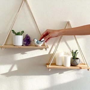 Peut inclure: Deux étagères suspendues en bois avec des supports en corde. L'étagère supérieure contient un pot en céramique blanc avec une plante, un cristal violet et un oiseau en céramique. L'étagère inférieure a des bougies et une petite plante en pot.