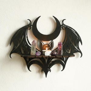 Könnte beinhalten: Schwarzes Holzregal in Fledermausform mit Mond und Fledermaus-Silhouette. Das Regal enthält Kristalle und eine brennende Kerze. Die Flügel haben aufwendige Ausschnitte, die eine gotische oder Hexen-Ästhetik erzeugen.