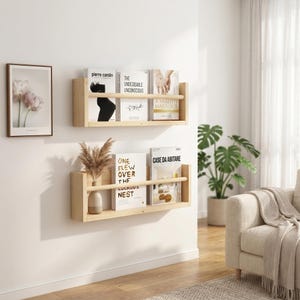 Peut inclure: Deux étagères flottantes en bois clair fixées au mur blanc, chacune contenant des livres et des objets décoratifs. L'étagère supérieure présente des livres avec des titres tels que "Pierre Cardin" et "A Healthy Marriage". L'étagère inférieure présente des livres et un vase.