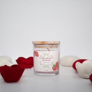 May include: A clear glass jar candle with a wooden lid, labelled "Strawberry Shortcake." The label features strawberry illustrations and decorative elements. Red and white fabric strawberry-shaped decorations surround the candle. The candle is 113g.