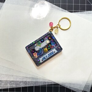 May include: A blue and silver glitter resin keychain shaped like a cassette tape with the text "HEY JUDE" on the front. The keychain is on top of a stack of white sheets of paper.