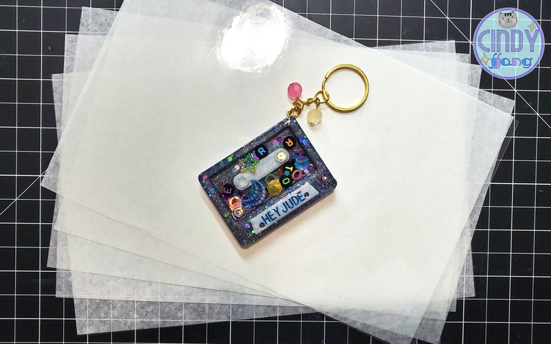 May include: A blue and silver glitter resin keychain shaped like a cassette tape with the text "HEY JUDE" on the front. The keychain is on top of a stack of white sheets of paper.