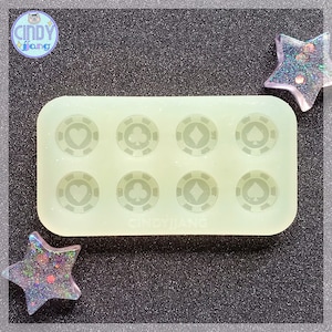 May include: A clear silicone mold with eight circular cavities. Each cavity features a raised design of a playing card suit: heart, club, diamond, and spade. The mold is on a black textured surface.