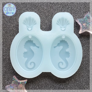 Puede incluir: Un molde de silicona azul claro para hacer pendientes. El molde presenta dos cavidades ovaladas, cada una con un diseño de caballito de mar. Encima de cada óvalo hay un diseño de concha. La marca "CINDY FANG" es visible.