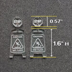 May include: Two clear acrylic warning signs with black text and graphics. The signs are 1.6 inches tall and 0.57 inches wide. The signs read "Caution Wet Floor" and feature a stick figure slipping on a wet surface. There is also a smaller sign that reads "Stop" in black text on a white background.