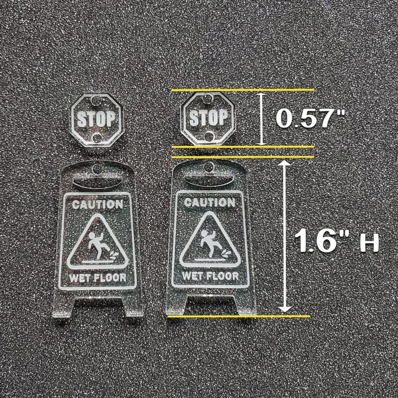 May include: Two clear acrylic warning signs with black text and graphics. The signs are 1.6 inches tall and 0.57 inches wide. The signs read "Caution Wet Floor" and feature a stick figure slipping on a wet surface. There is also a smaller sign that reads "Stop" in black text on a white background.