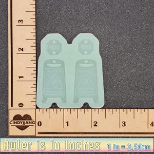 May include: Two clear silicone molds for making resin crafts. Each mold is shaped like a caution sign with the words "Caution" and "Slippery When Wet" in a black font. The molds are 1.5 inches tall and 1 inch wide.