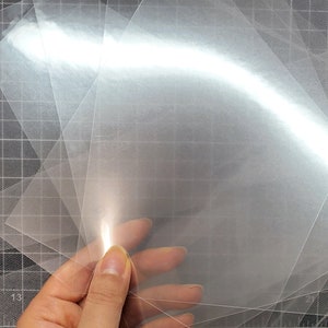 May include: A stack of clear plastic sheets, likely for crafting or scrapbooking, on a black grid surface.