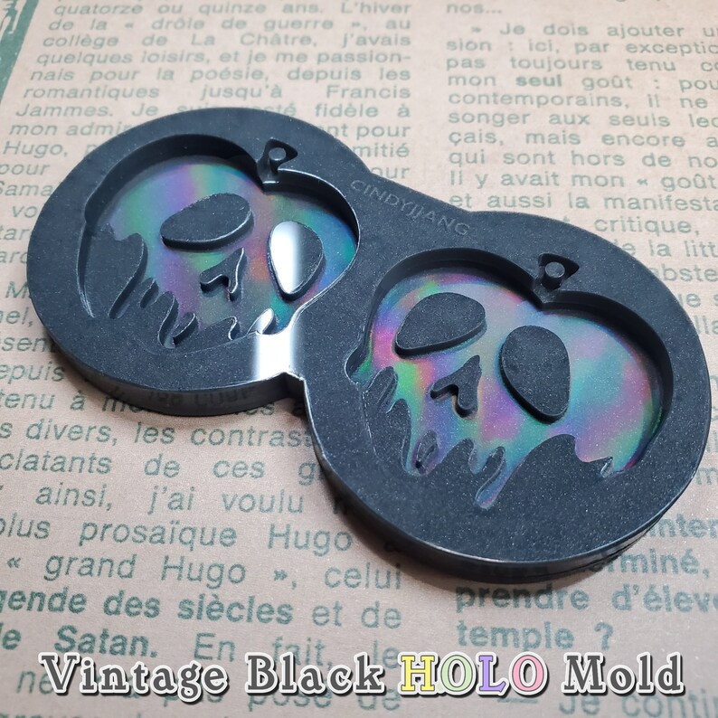 May include: Black silicone mold with two cavities shaped like an apple with a skull design. The mold is designed to create resin crafts with a holographic finish. The mold is on a textured surface with text that reads "Vintage Black HOLO Mold".