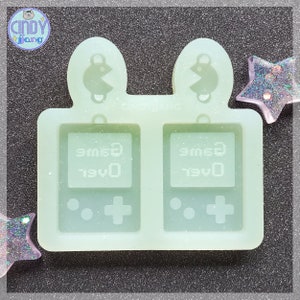 May include: A clear silicone mold for making earrings. The mold has two sections, each with a design of a retro video game console with the text "game over". The top of each section has a Pac-Man design.