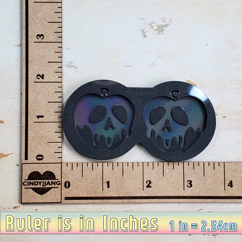 May include: A black silicone mold for making resin earrings. The mold features two poison apple designs with iridescent green, blue, and purple accents. The mold is 2 inches wide.
