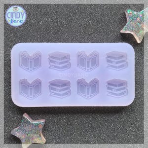 May include: A clear silicone mold with six different book shapes. The mold is designed to create resin book charms. The mold has a light purple hue.