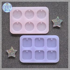 Könnte beinhalten: Zwei Silikonformen, eine rosa mit sechs kreisförmigen Hohlräumen und eine lila mit sechs quadratischen Hohlräumen. Zwei glitzernde sternförmige Harzstücke sind ebenfalls vorhanden. Die Formen dienen zur Herstellung von Schmuck oder kleinen Dekorationsartikeln.