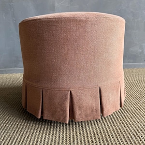 May include: A round, upholstered ottoman in a muted reddish-brown hue. The fabric has a textured appearance, and the base features a pleated skirt detail. The ottoman is a versatile piece of furniture.