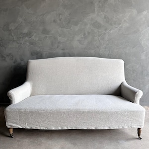 May include: A white linen sofa with a curved back and rolled arms. The sofa has wooden legs with casters and is sitting on a gray concrete floor.