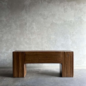 May include: A rectangular wooden bench with a natural finish. The bench has a simple, blocky design with two sturdy legs. The wood grain is visible, adding texture and warmth to the piece. The bench is approximately 48 inches long.