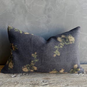 May include: A rectangular decorative pillow with a dark blue background and a floral pattern in shades of green and beige. The pillow is made of a textured fabric and is placed on a wooden surface.