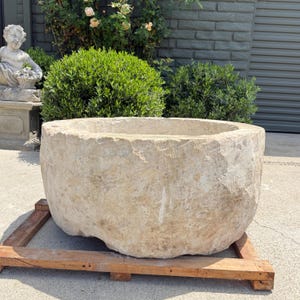 May include: A large, weathered stone basin with a rough, textured surface. The basin is a light beige color and sits on a wooden pallet. The basin has a wide, open top and a rounded shape. The background includes greenery and a statue.