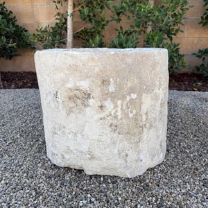 May include: A weathered, cylindrical stone planter with a rough, textured surface. The planter is a light beige color with darker patches and sits on a bed of small gray pebbles. The shape is slightly tapered.