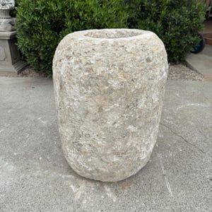 May include: A weathered, cylindrical stone planter with a wide opening at the top. The planter has a rough, textured surface and a light gray color. The outdoor decor item is suitable for a garden or patio.