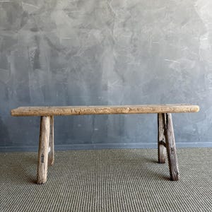 May include: A rustic, wooden bench with a natural, weathered appearance. The bench has a long, rectangular seat supported by four sturdy, angled legs. The wood has visible grain and knots, adding to its aged charm. The bench is set against a gray wall and a striped rug.