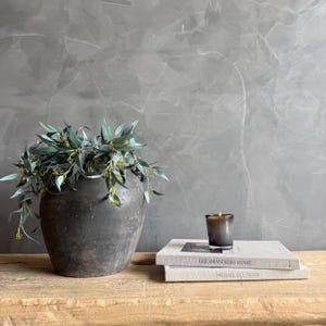 May include: A rustic, dark gray ceramic vase filled with green and yellow leafy branches. A small lit candle sits atop two books with the titles "LES AMANDIERS HOME" and "MICHAEL DEL PIERO" on a wooden surface. The background is a textured gray wall.