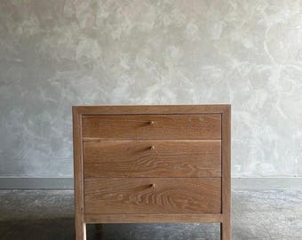 May include: A three-drawer wooden nightstand with a light wood finish. The drawers have brass pulls and are slightly recessed.