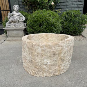 May include: A large, weathered stone planter with a cylindrical shape. The planter has a rough, textured surface and a wide opening at the top. The color is a light beige with variations in tone. The planter is outdoors, near a statue and greenery.