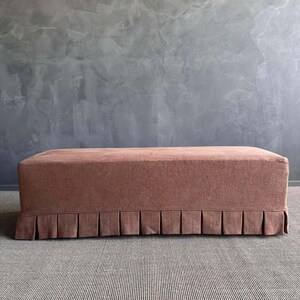 May include: A rectangular, upholstered ottoman in a muted rose color. The ottoman features a pleated skirt along the bottom edge. It is placed on a gray and white striped rug, with a textured gray wall in the background.