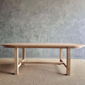 May include: A light wood oval dining table with a simple, modern design. The table has a rectangular base with four legs and a crossbar.