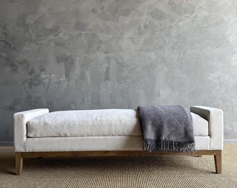 May include: A light-colored upholstered bench with a wooden frame and legs. A gray blanket with fringe is draped over the bench. The bench is on a striped rug, and the background is a textured gray wall.