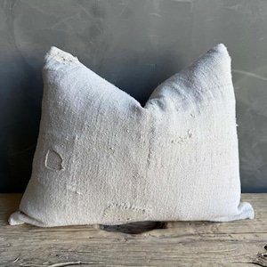 May include: A light gray, textured pillow with a distressed appearance. The pillow has a rectangular shape with a pointed top and visible patches. The pillow is made of a woven fabric and rests on a wooden surface.