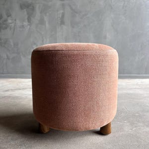 May include: A round, pink upholstered ottoman with wooden legs. The ottoman is sitting on a gray concrete floor.