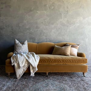 May include: A mustard yellow velvet sofa with three decorative pillows and a cream-colored throw blanket. The sofa has a curved back and wooden legs. The pillows are in shades of brown and white. The background is a textured gray wall.