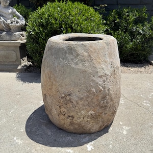May include: A large, weathered, beige stone planter with a wide opening. The cylindrical pot has a rough, textured surface and sits on a concrete surface. The planter is outdoors, with greenery in the background.