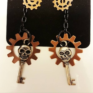 May include: A pair of silver skull and key earrings with copper gears and black chains. The earrings are on a black card with the text "By Ravenseed".