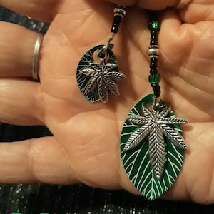 May include: A pair of green and silver earrings with a marijuana leaf design. The earrings are made with black beads and silver charms. The earrings are hanging from a person's hand. By Ravenseed.