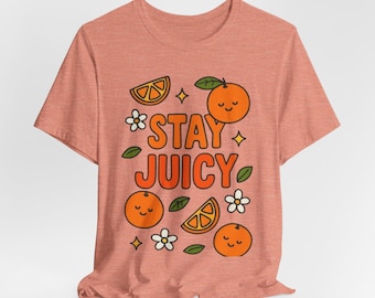 Könnte beinhalten: Korallfarbenes T-Shirt mit dem Schriftzug "STAY JUICY" in Orange. Das Design umfasst Cartoon-Orangen, Orangenscheiben, weiße Blumen, grüne Blätter und Sternakzente. Das T-Shirt hat einen Rundhalsausschnitt und kurze Ärmel.
