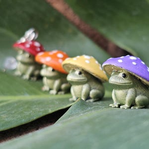 Puede incluir: Cuatro peque&ntilde;as figuras de rana con sombreros de seta en rojo, naranja, amarillo y morado con lunares blancos. Las ranas son verdes y est&aacute;n sentadas sobre una gran hoja verde. Los sombreros de seta est&aacute;n pintados con un acabado brillante.