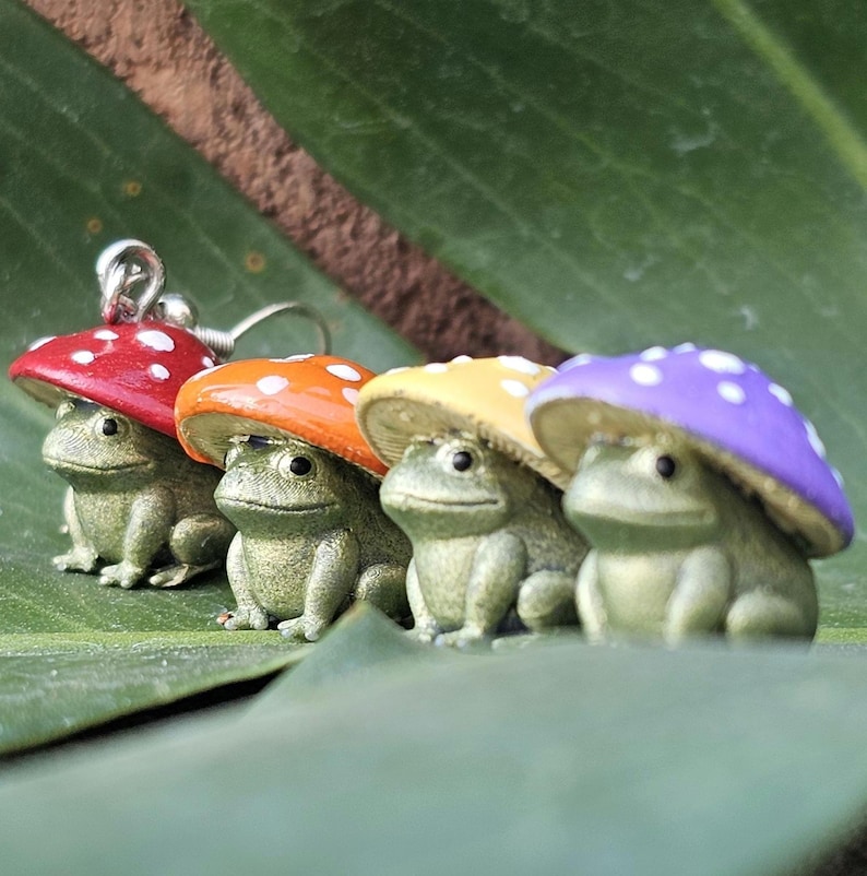 Puede incluir: Cuatro pendientes de rana verde, cada uno con una gorra de hongo en rojo, naranja, amarillo y morado con lunares blancos. Las ranas sonr&iacute;en. Los pendientes est&aacute;n sobre una hoja verde.