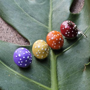 Puede incluir: Cuatro adornos con forma de hongo con lunares blancos descansan sobre una gran hoja verde. Los hongos son morados, amarillos, naranjas y rojos. Un hongo rojo es un pendiente con un gancho plateado. La hoja tiene venas prominentes.