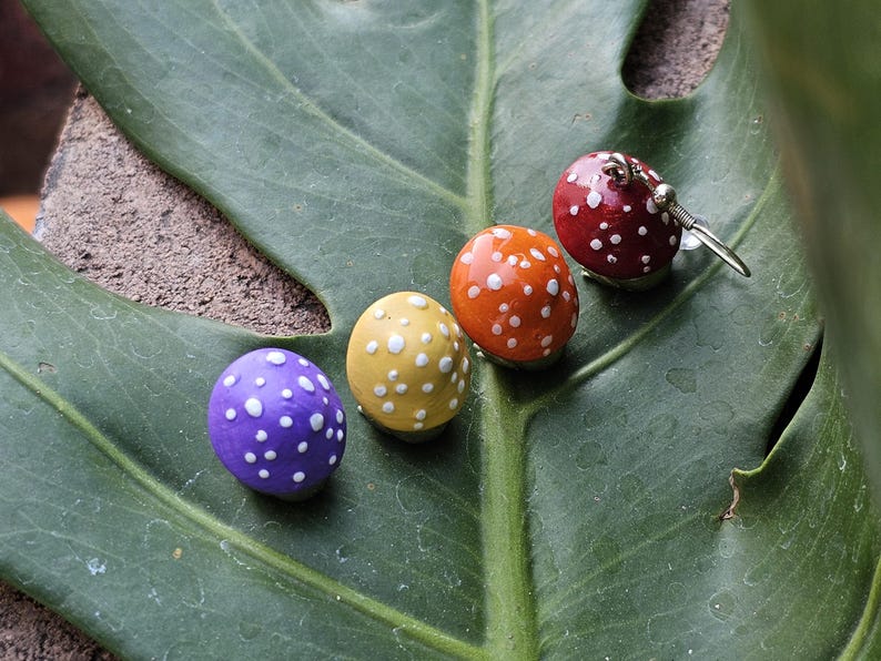 Puede incluir: Cuatro adornos con forma de hongo con lunares blancos descansan sobre una gran hoja verde. Los hongos son morados, amarillos, naranjas y rojos. Un hongo rojo es un pendiente con un gancho plateado. La hoja tiene venas prominentes.