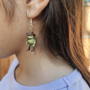 May include: A single earring featuring a small, green frog figurine wearing a brown cowboy hat and boots. The frog hangs from a silver-colored fish hook earring.