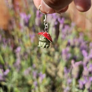 Puede incluir: Un pendiente caprichoso con una peque&ntilde;a rana verde que lleva un gorro de seta roja con lunares blancos. La rana est&aacute; suspendida de un gancho plateado. El fondo es un campo borroso de flores moradas.