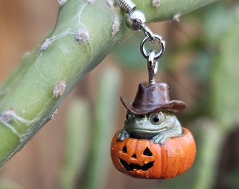May include: A silver-toned earring featuring a small green frog wearing a brown cowboy hat, sitting inside a carved orange jack-o'-lantern pumpkin. The charm is attached to a standard metal earring hook, showcasing detailed textures on the pumpkin.