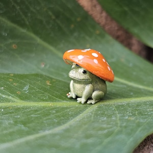 Puede incluir: Una peque&ntilde;a figura de rana verde con un sombrero de hongo naranja con lunares blancos, sentada sobre una gran hoja verde. La figura es una decoraci&oacute;n de jard&iacute;n caprichosa.