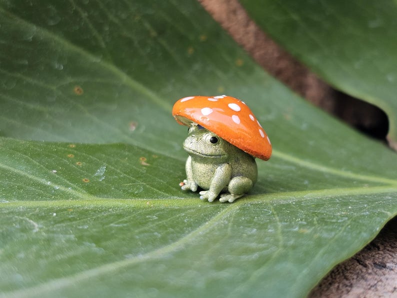 Puede incluir: Una peque&ntilde;a figura de rana verde con un sombrero de hongo naranja con lunares blancos, sentada sobre una gran hoja verde. La figura es una decoraci&oacute;n de jard&iacute;n caprichosa.