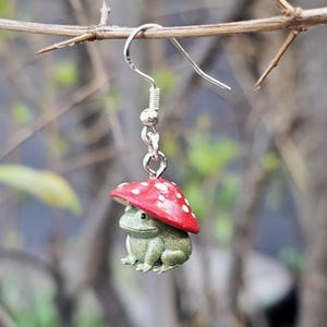 May include: A whimsical silver earring featuring a small green frog wearing a red mushroom cap with white spots. The frog is detailed with textured skin and sits beneath the mushroom. The earring hangs from a silver hook.