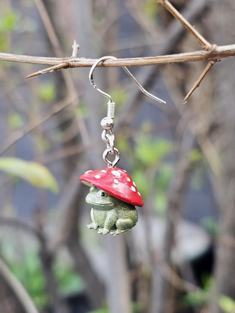 Puede incluir: Un pendiente de plata caprichoso con una peque&ntilde;a rana verde que lleva una gorra de hongo rojo con lunares blancos. La rana tiene la piel texturizada y el pendiente cuelga de un gancho de plata.