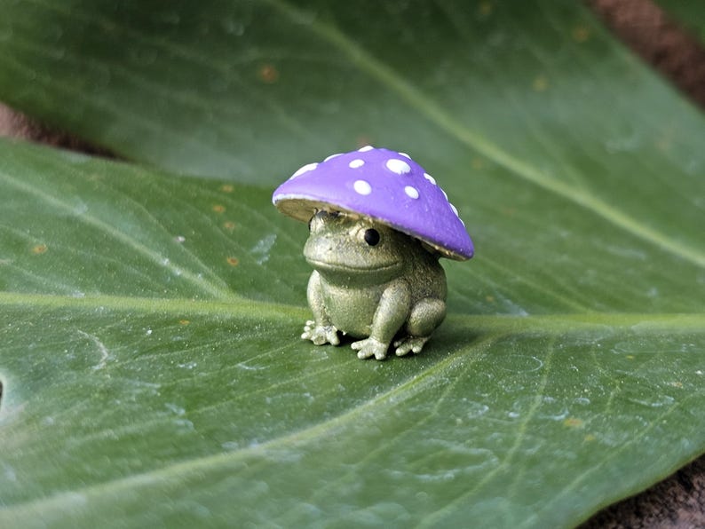Puede incluir: Una peque&ntilde;a figura de rana verde con una gorra de hongo morado con lunares blancos. La rana est&aacute; sentada en una hoja grande y verde. La figura tiene una est&eacute;tica caprichosa de cuento de hadas.